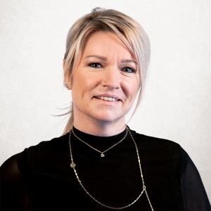 Porträt einer Frau mit blondem Haar in einem Zopf und schwarzes Oberteil mit Rollkragen vor hellem Hintergrund, schaut lächelnd in die Kamera.