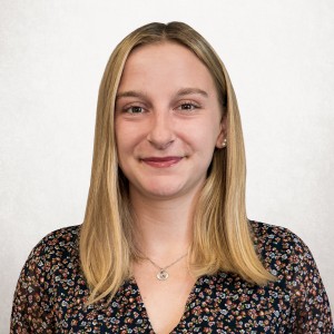 Porträt einer Frau mit blondem Haar und floraler Bluse vor hellem Hintergrund, schaut lächelnd in die Kamera.